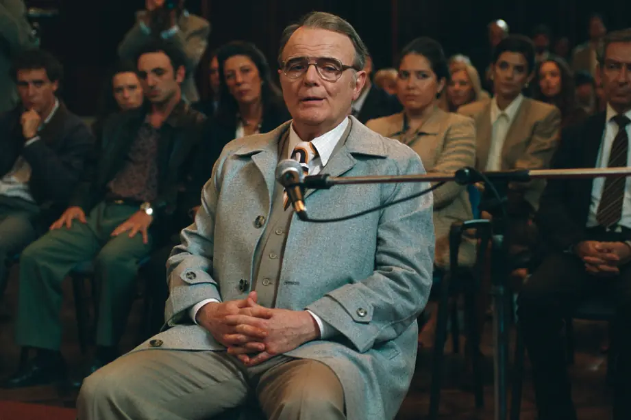 Fotograma de la película Barreda. Luis Machín, en el papel de Ricardo Barreda, declara sentado ante el micrófono en la sala de audiencias, con el público detrás.